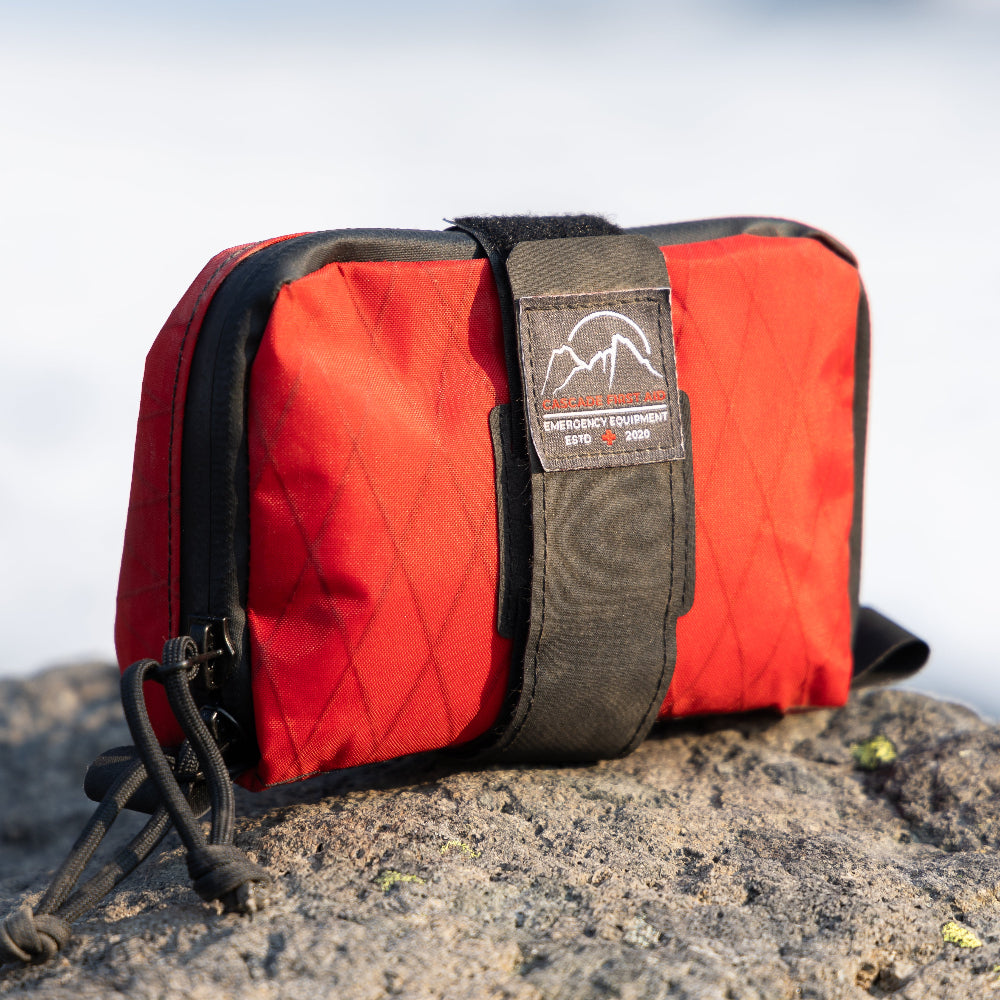 Traverse lightweight backcountry first aid kit in red, sitting on the rocks on top of a mountain during a backpacking trip in central Oregon. From Cascade First Aid, photo by @chrisinthewater